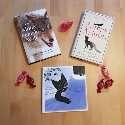 Three books on a table, Aesop's Animals, The Alpha Female Wolf, and PS I Love You More Than Tuna, along with a red bird ornament and some red sweets